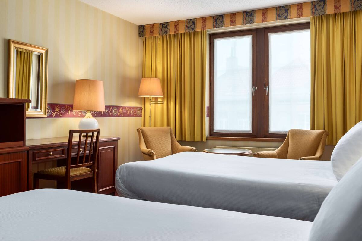 A hotel room with two beds, a desk, a chair, a lamp, and a window with curtains.