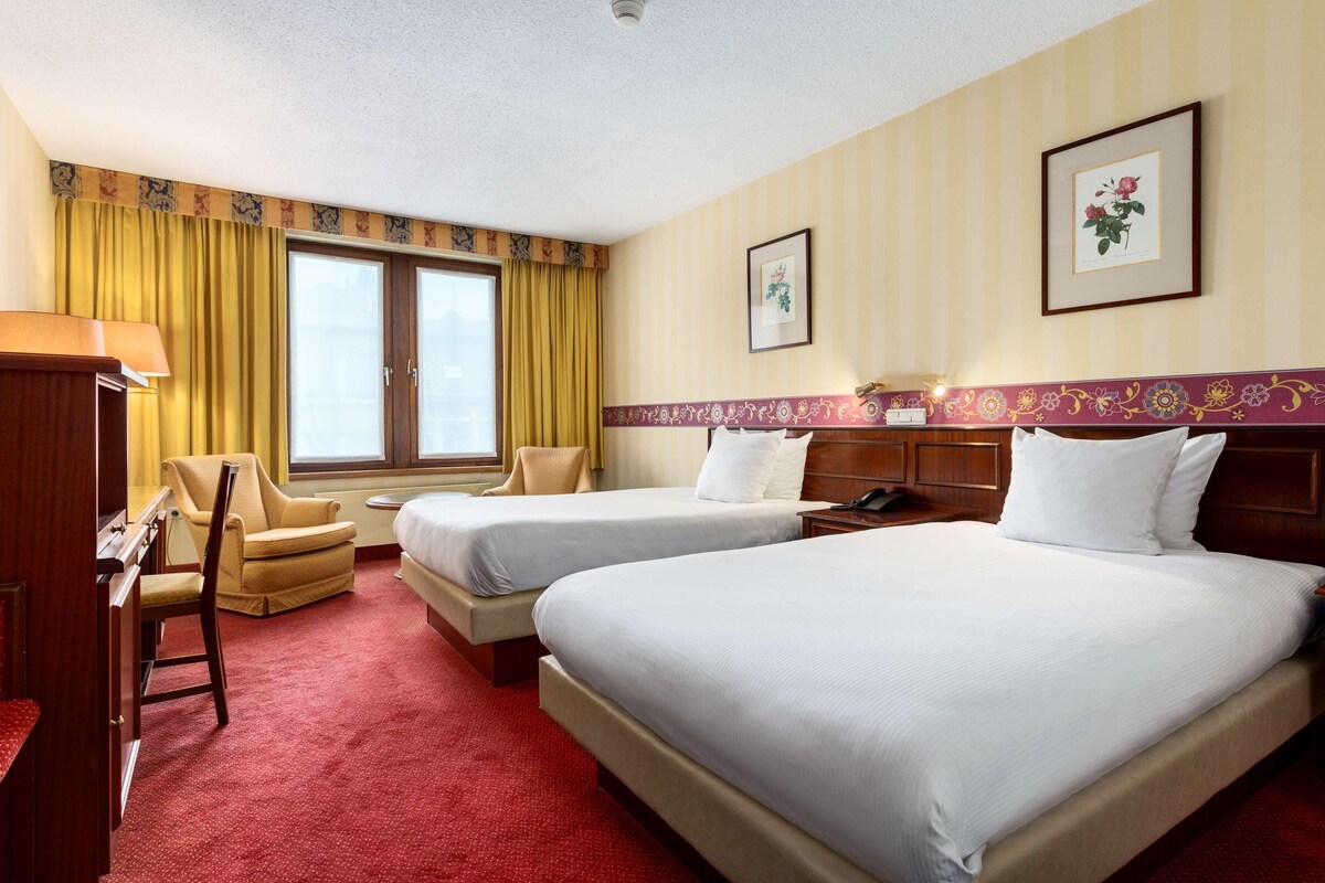 A hotel room with two beds, a desk, a chair, and a window with curtains.