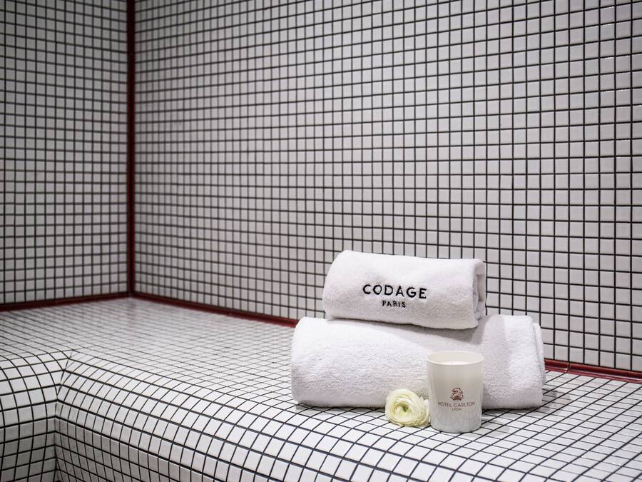 A bathroom with a grid-patterned tile floor and walls, a white towel with "CODAGE PARIS" text, and a white candle with "HOTEL CAPTON LONDON" text.