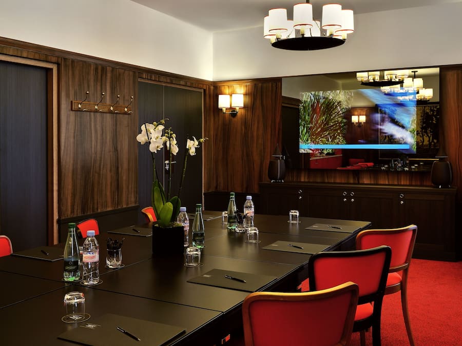 A conference room with a long table, red chairs, a large flat-screen TV, and two chandeliers.