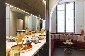 A modern bathroom with a large mirror, a countertop with various glass containers, and a red sofa in the adjacent living area. - Carlton Hotel Lyon - MGallery Collection (Lyon)