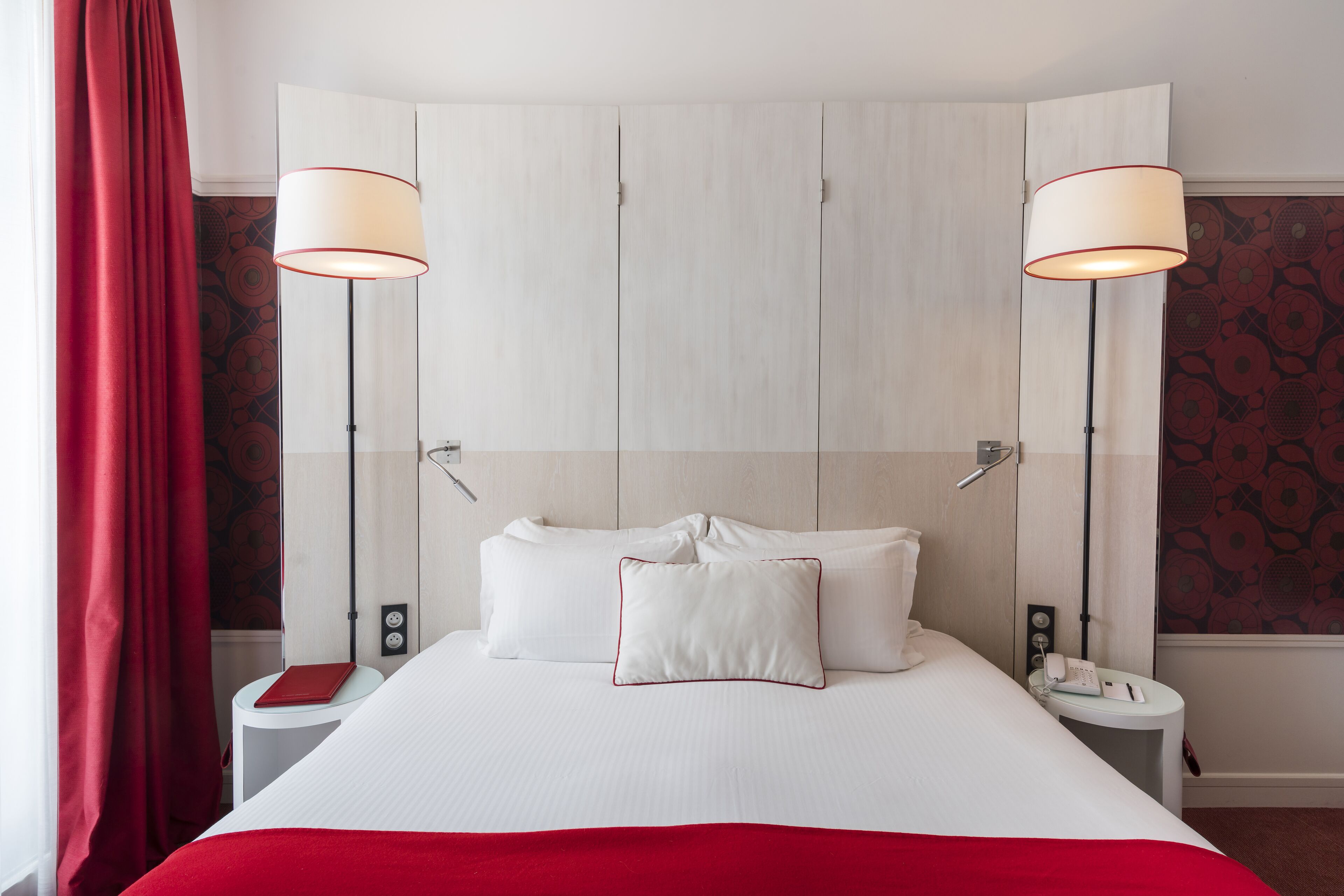 A hotel room with a large bed, two bedside lamps, a bedside telephone, and a red curtain.