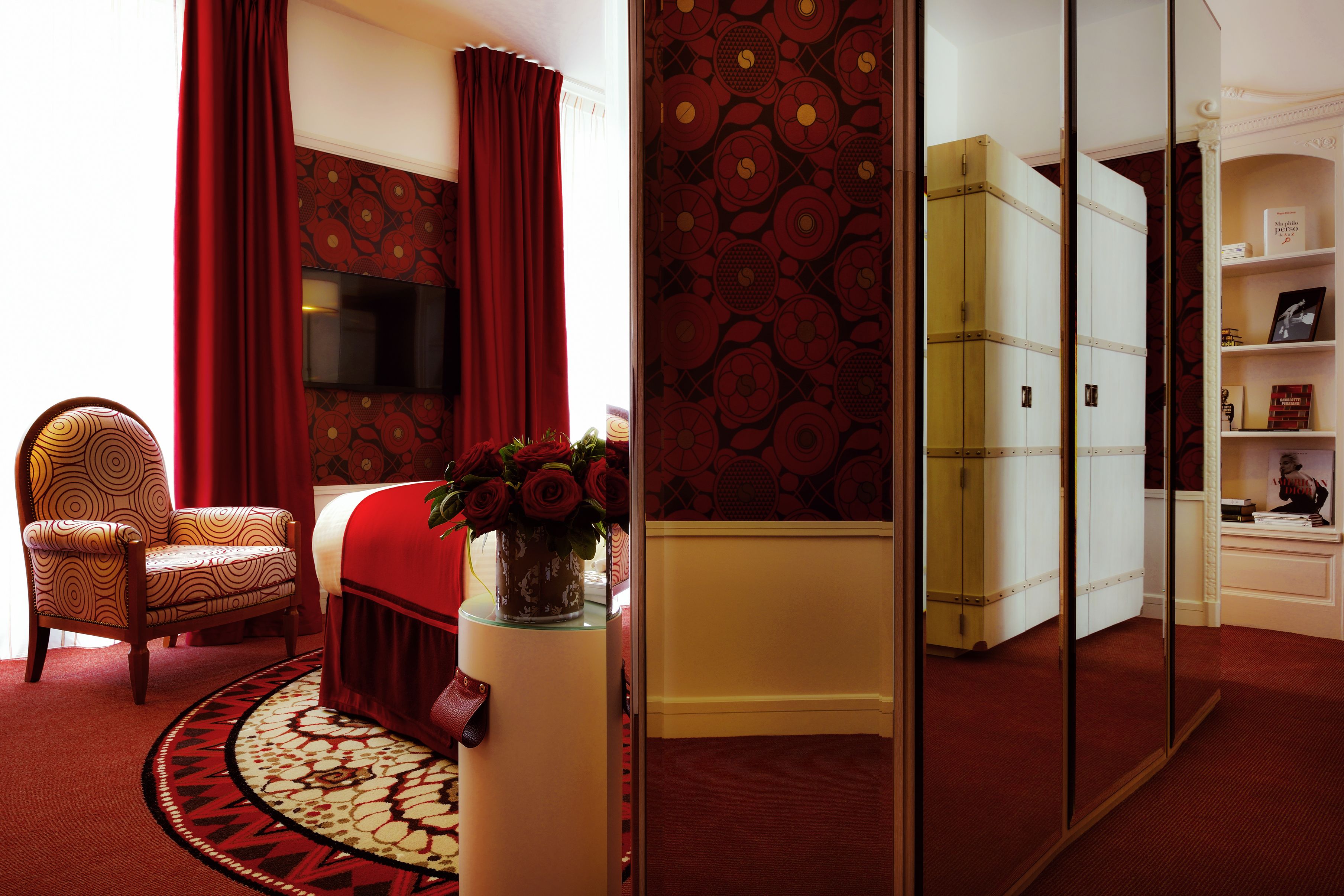 Una habitación con alfombra roja, un sillón estampado, un sofá rojo, una mesa de vidrio con un florero y un televisor, además de un armario con espejo.