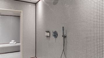 Une salle de bain moderne avec un mur recouvert de carreaux disposés en motif quadrillé, un banc avec des serviettes et un espace douche doté d’un pommeau de douche à main.