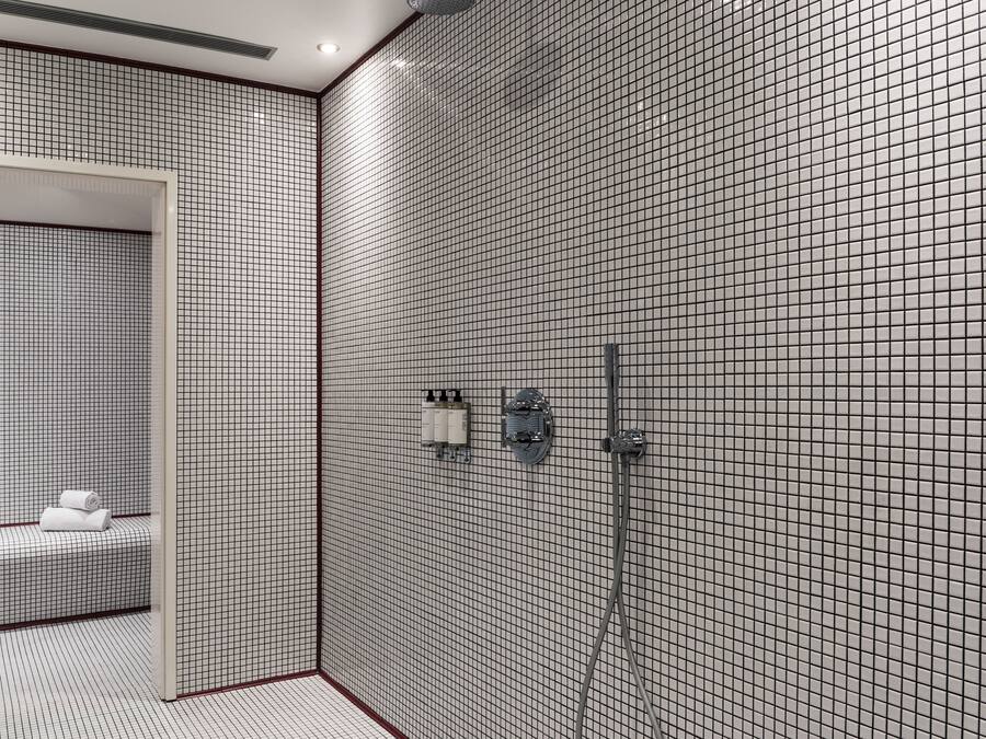 Un baño moderno con pared de azulejos formando un patrón de cuadrícula, un banco con toallas y una zona de ducha con cabezal de ducha de mano.