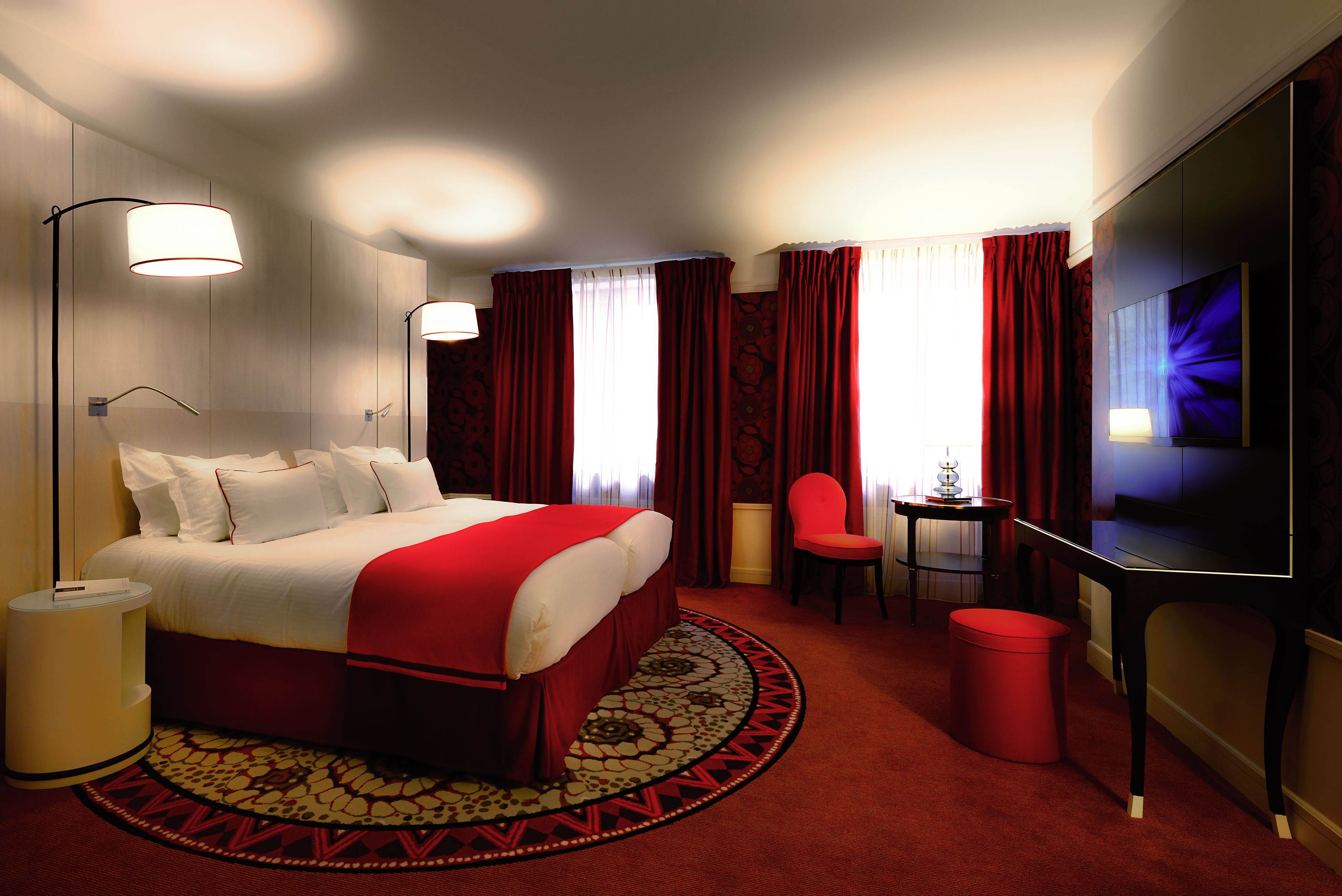 A hotel room with a large bed, a desk, a chair, a TV, and a red carpet.