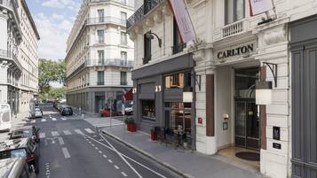 A street view with a Carlton hotel entrance, a crosswalk, parked cars, and a building with a flag.