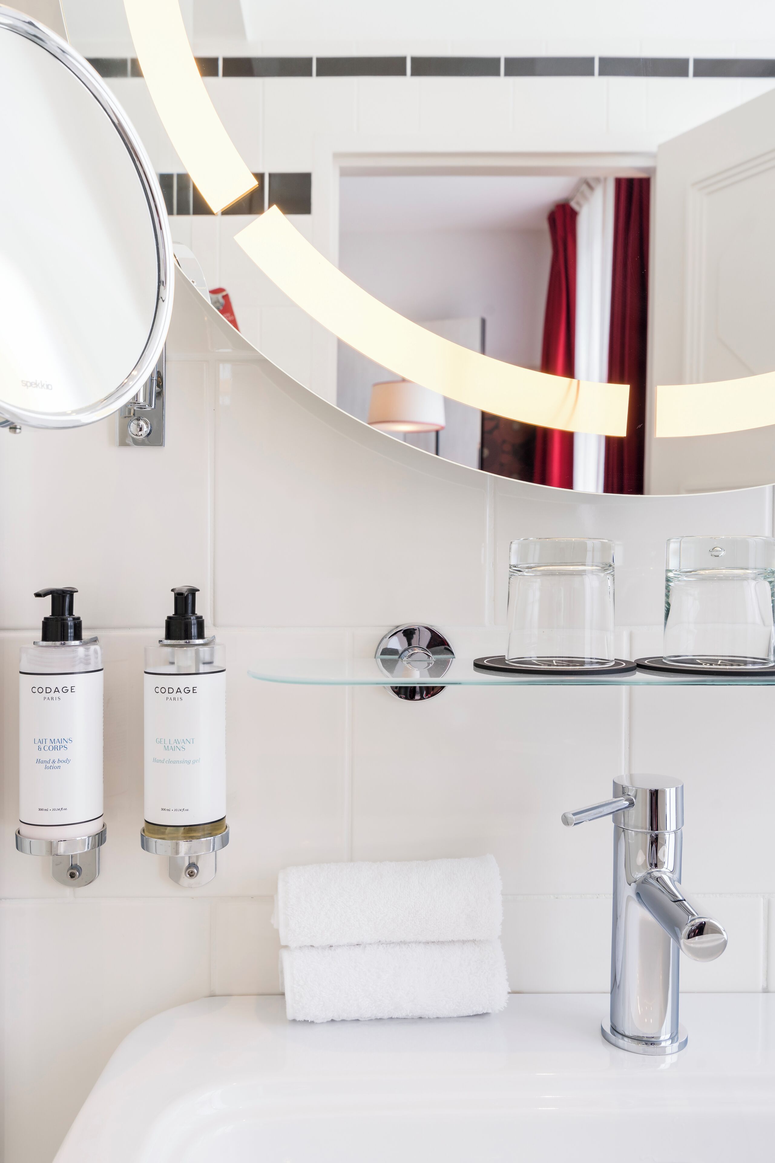 A modern bathroom with a white sink, a mirror, two hand soap dispensers, a glass shelf with two glasses, and a towel rack with two towels.