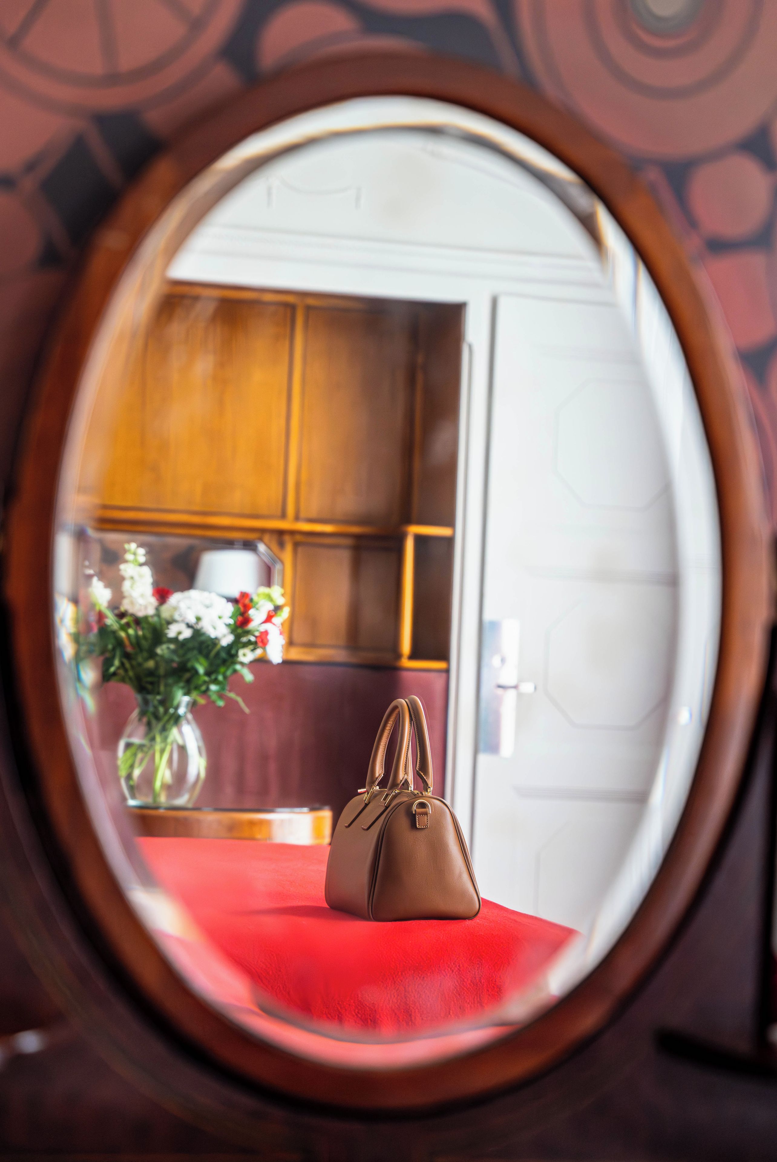 A round mirror reflecting a room with a red upholstered chair, a brown handbag, and a vase with flowers.