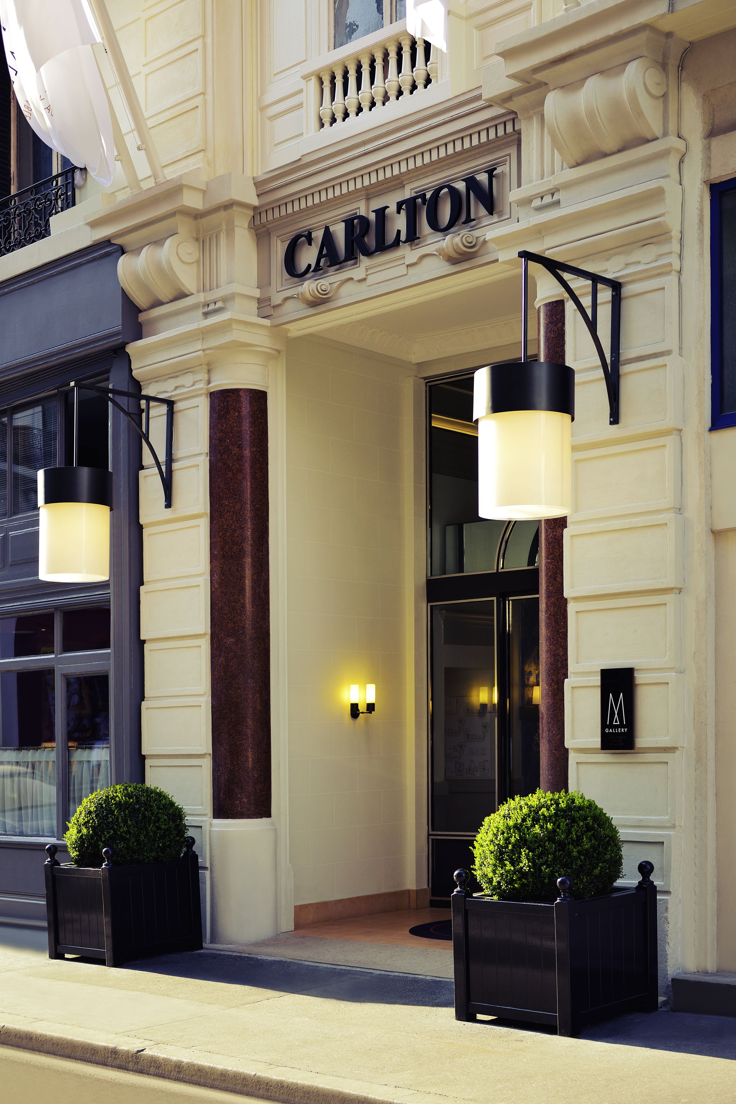 A hotel entrance with a sign reading "CARLTON," two black planters with green bushes, and a modern light fixture.