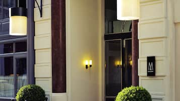 A hotel entrance with a sign reading "CARLTON," two black planters with green bushes, and a modern light fixture.