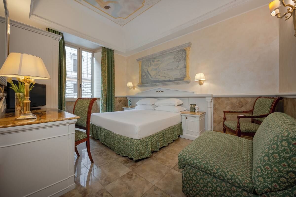 Ein Hotelzimmer mit Bett, Schreibtisch mit Lampe, Sessel, Nachttisch und einem Bild an der Wand.