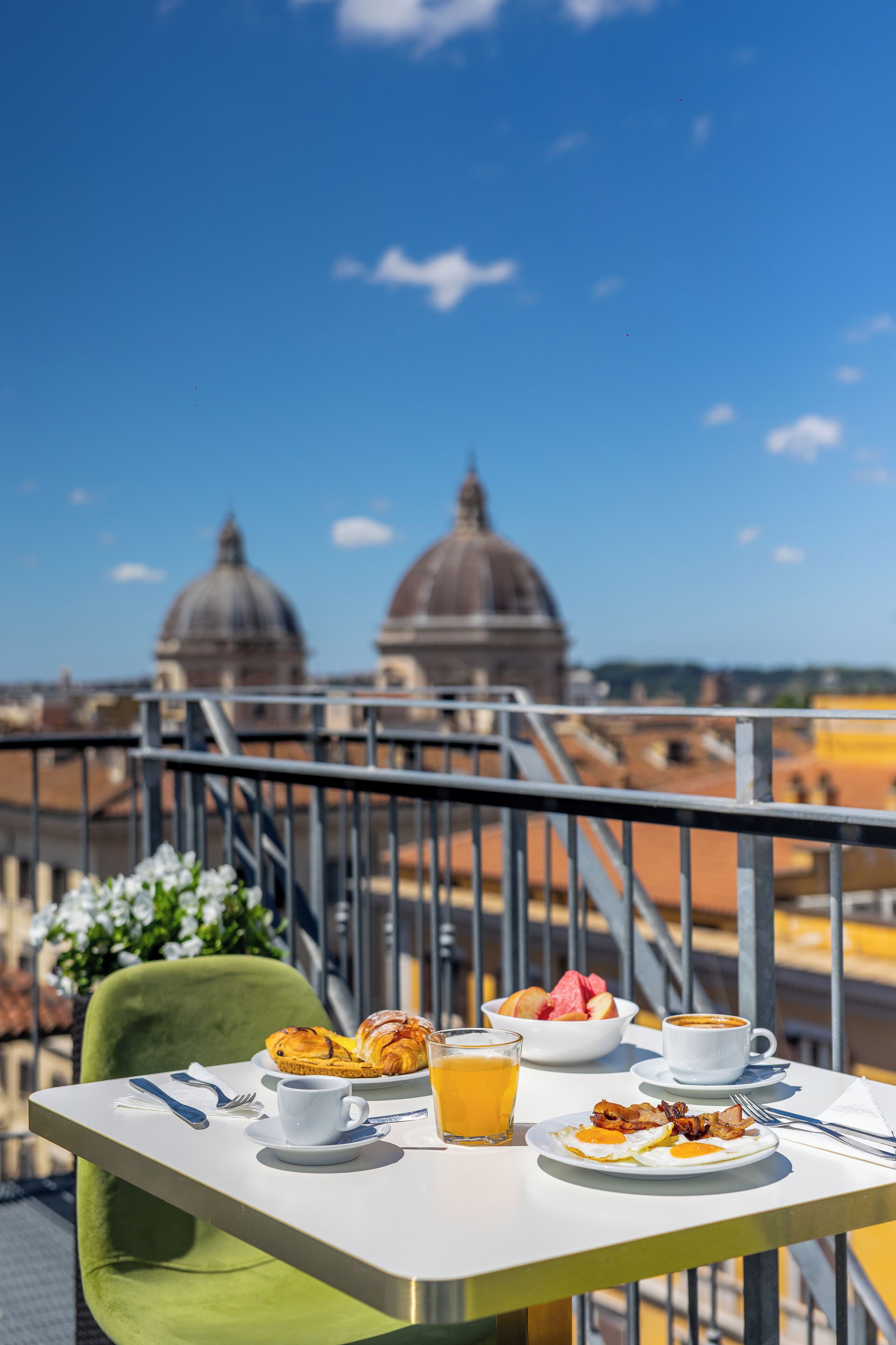 A breakfast table with croissants, eggs, and orange juice, overlooking a cityscape with domed buildings.