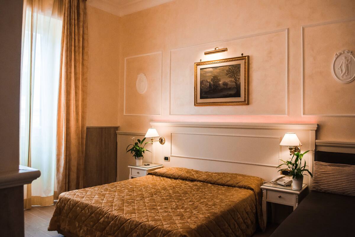 Ein Hotelzimmer mit einem großen Bett, zwei Nachttischen, einer Lampe, einem gerahmten Bild an der Wand und einem Fenster mit Vorhängen.