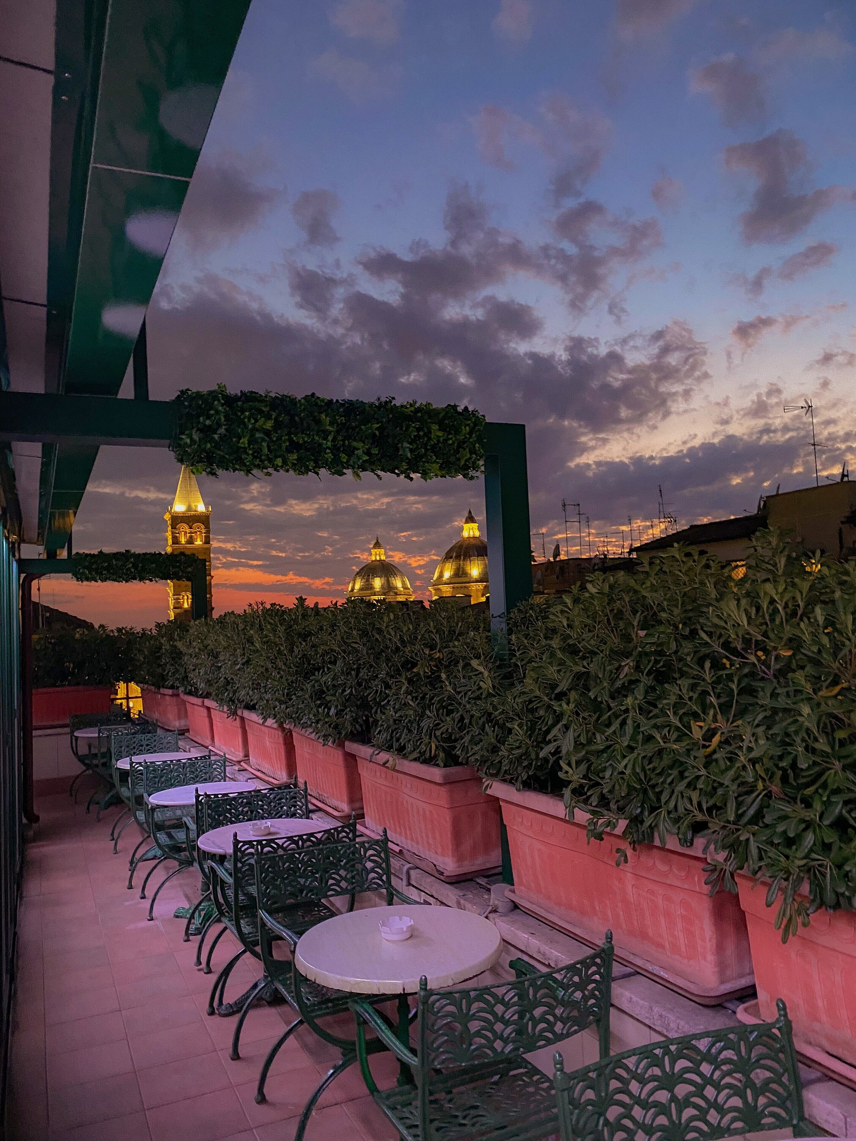 Un espace extérieur aménagé avec des tables et des chaises, une pergola couverte de verdure et une vue sur des bâtiments à coupoles se détachant sur un ciel crépusculaire.