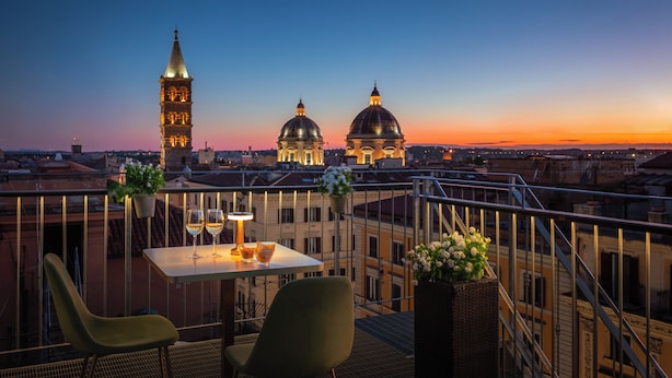 A terrace with a table, chairs, and a view of historic buildings at sunset. - Hotel Palladium Palace (Rome)