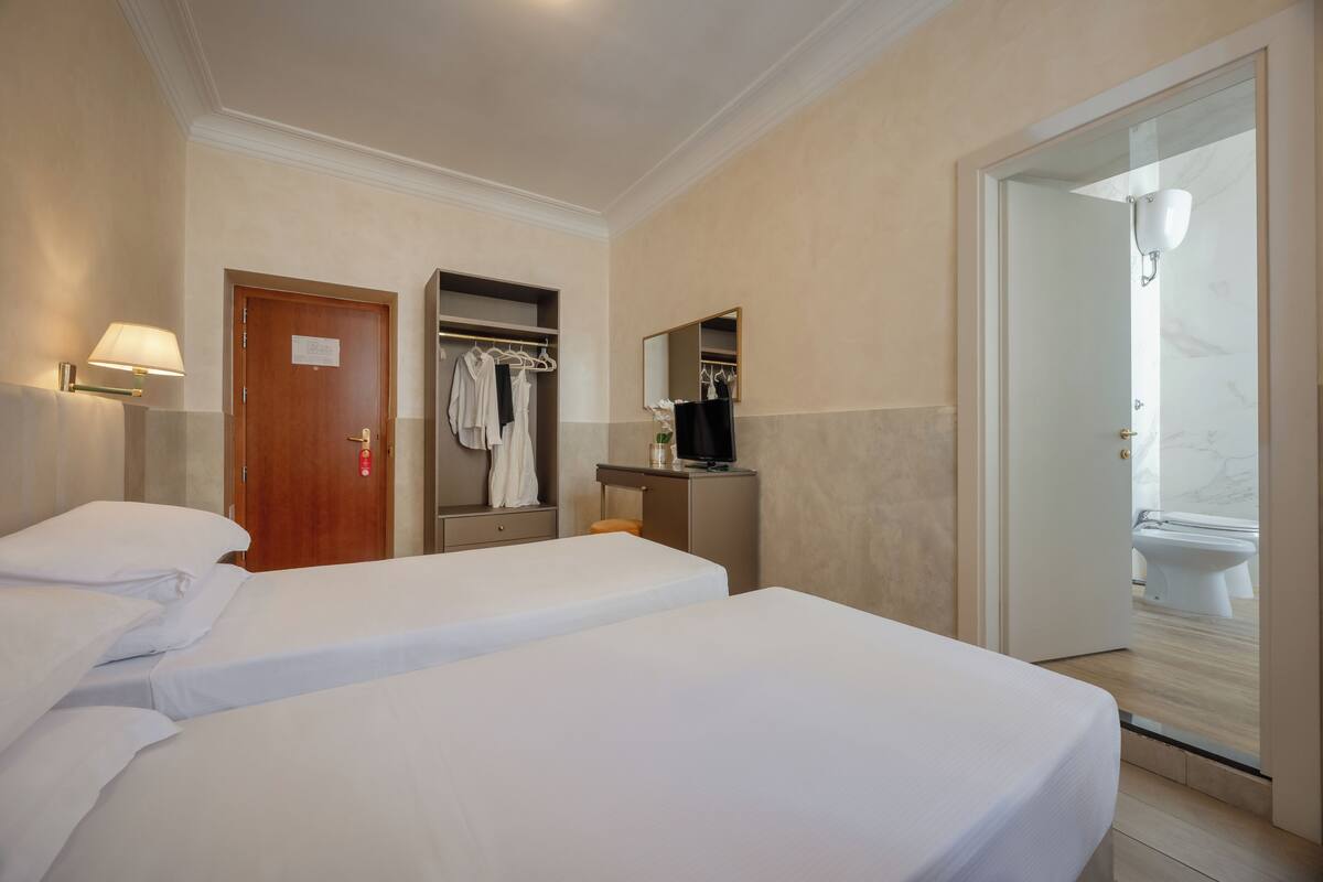 Ein Hotelzimmer mit zwei Betten, einem Kleiderschrank, einem Spiegel, einem Fernseher und einem durch eine offene Tür einsehbaren Badezimmer.