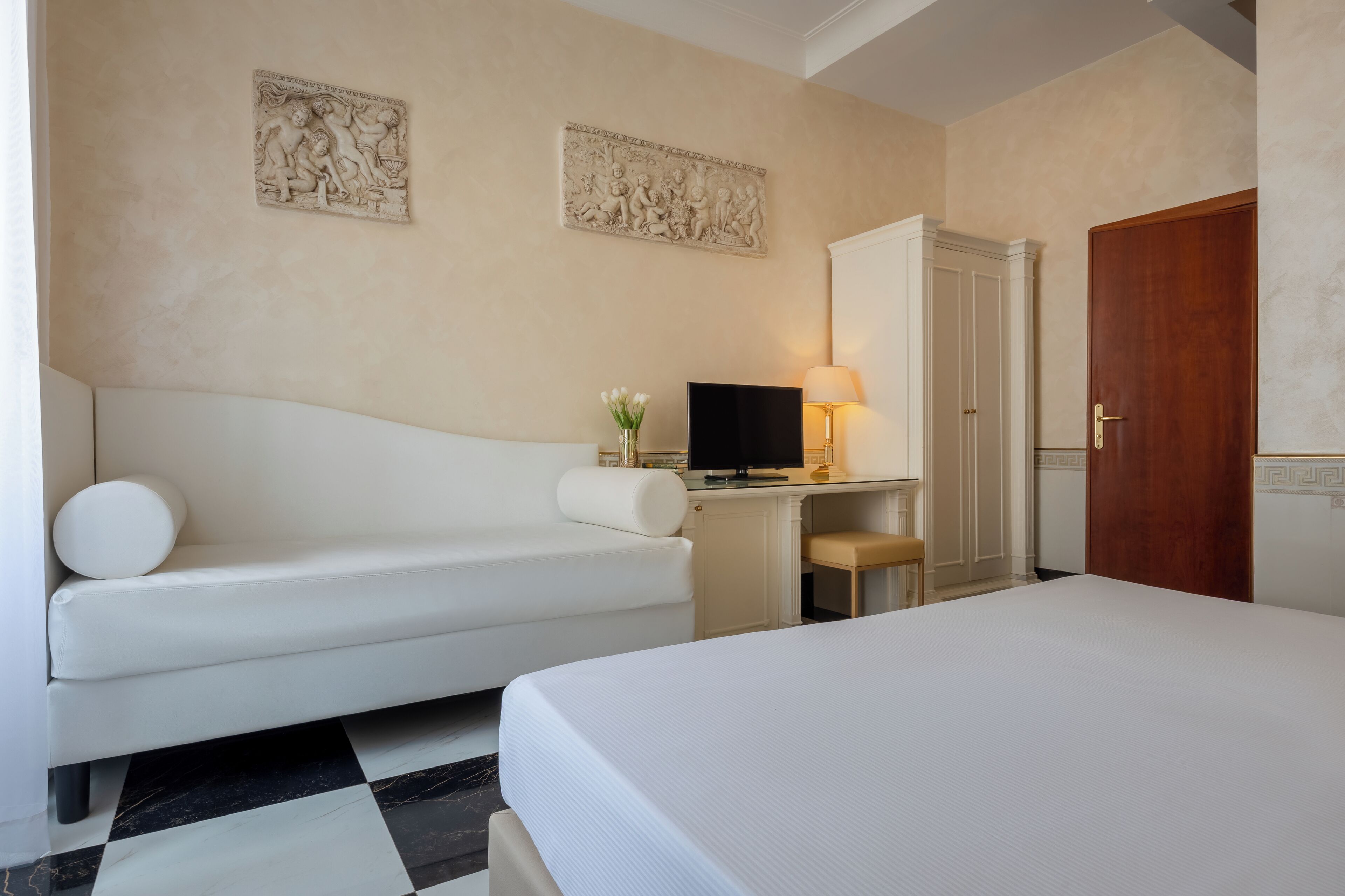 A hotel room with a bed, a sofa, a desk with a TV, a lamp, and two wall-mounted artworks.
