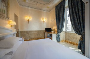 Et hotelværelse med en stor seng, et skrivebord med en computer, to væglamper og et vindue med gardiner.
