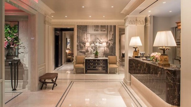 Um hall de hotel luxuoso com piso de mármore, balcão de recepção, área de estar com sofá e poltronas, e um mural decorativo na parede.