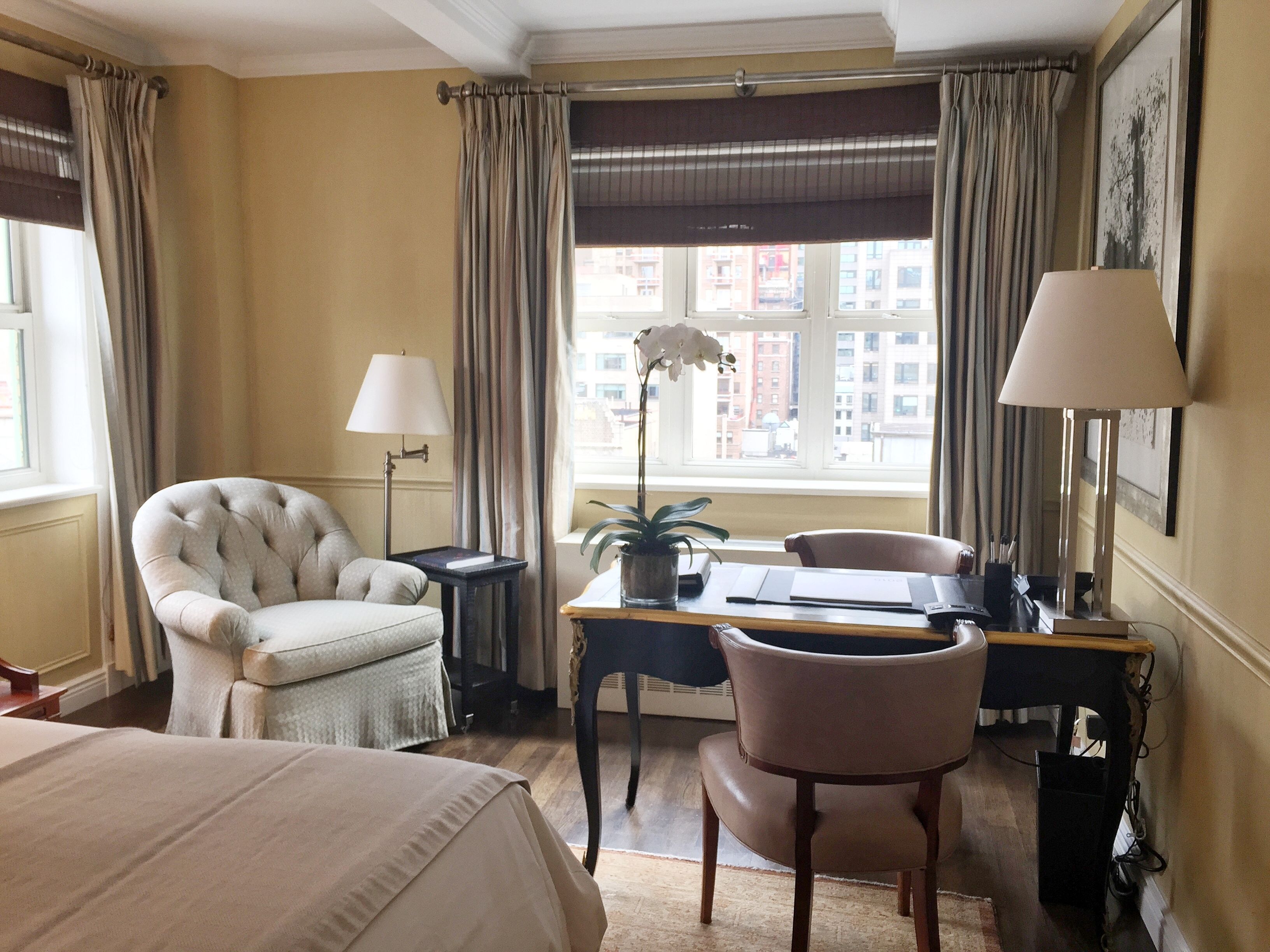 Une chambre d’hôtel avec un lit, un bureau et une chaise, une lampe, une plante en pot et une fenêtre avec des rideaux.