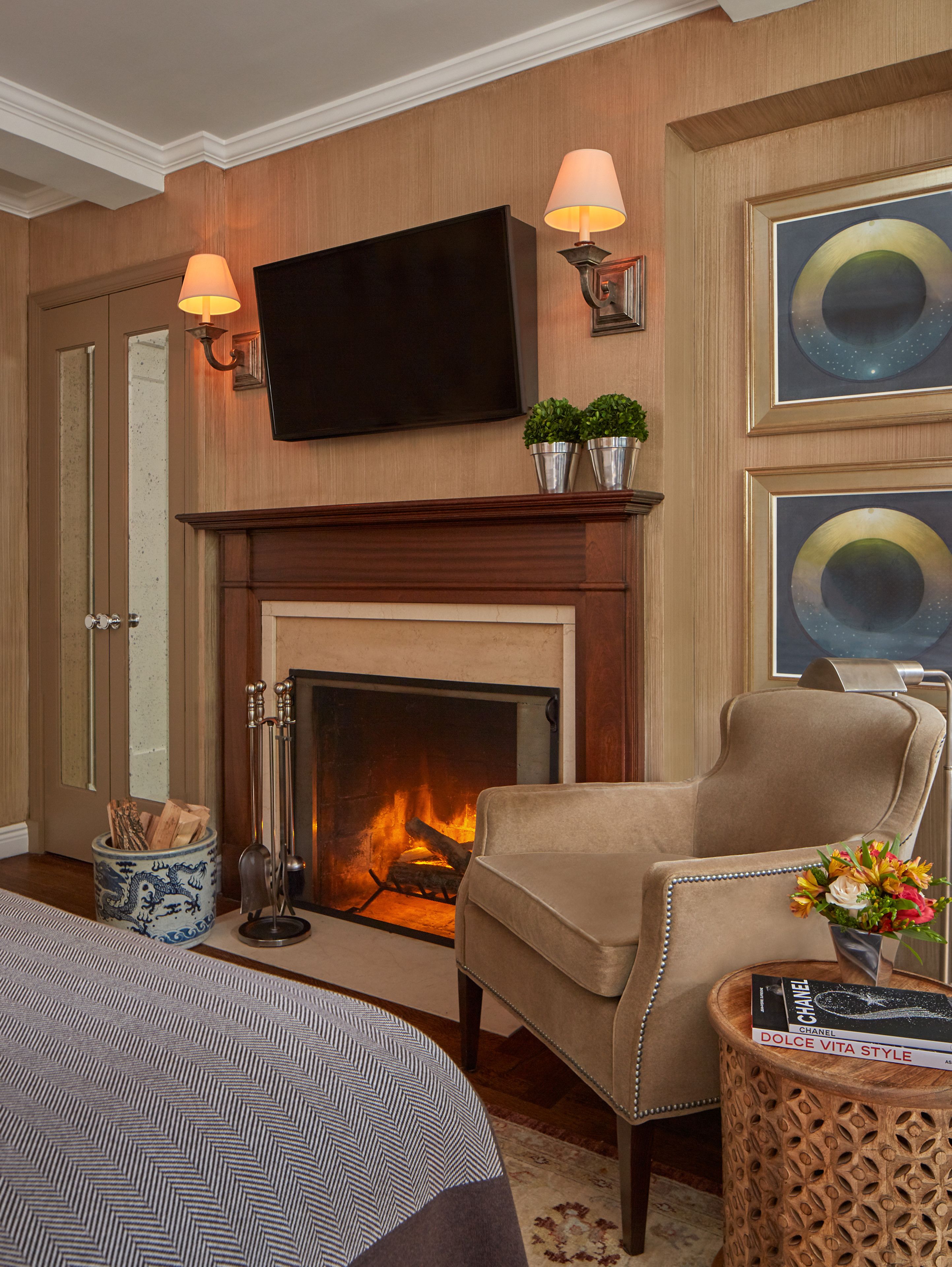 Habitación de hotel con chimenea, un sillón cómodo, un televisor montado sobre la chimenea y una mesita de noche con un libro y un jarrón de flores.