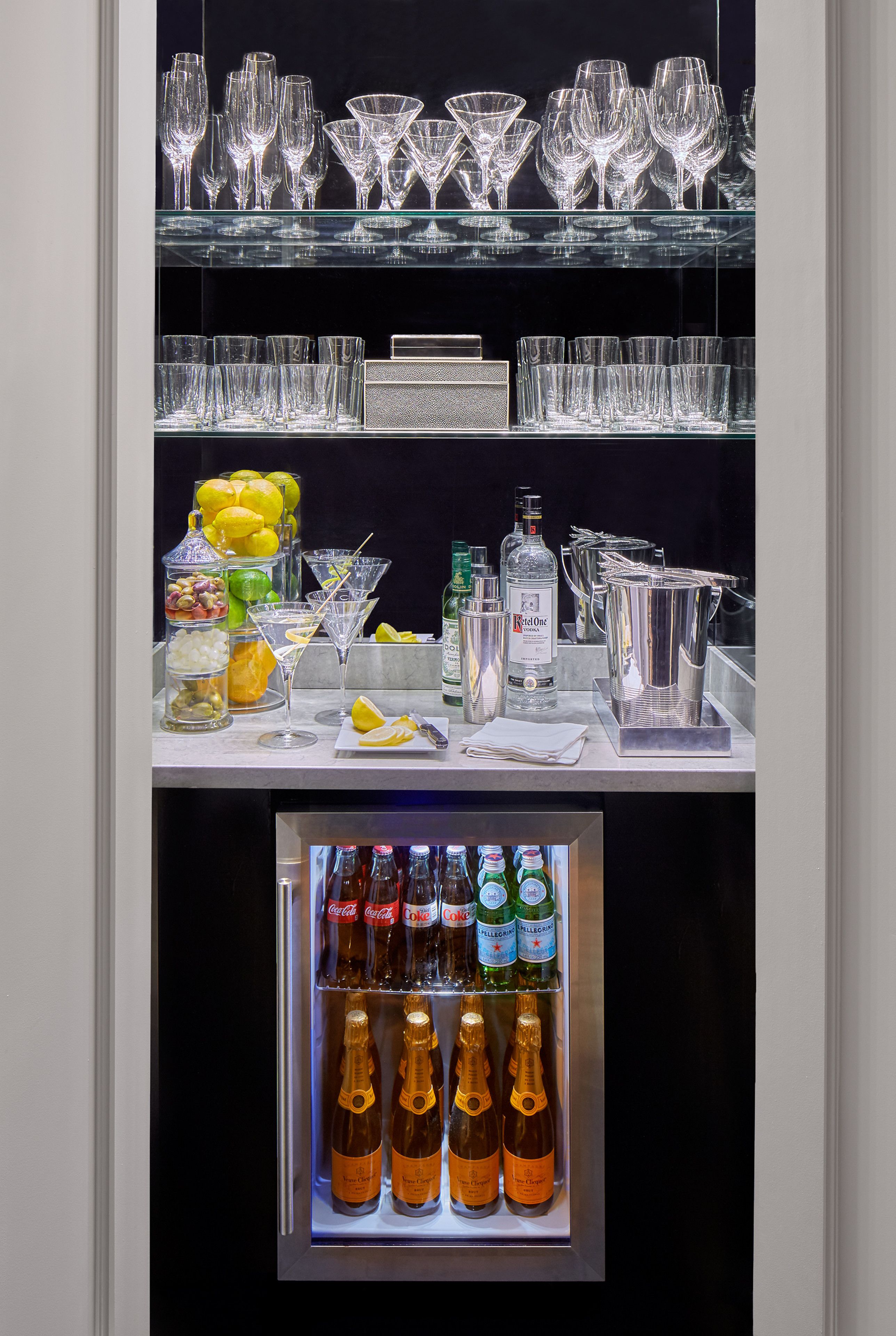 Zona de bar con diferentes copas, una variedad de botellas y un refrigerador lleno de bebidas.