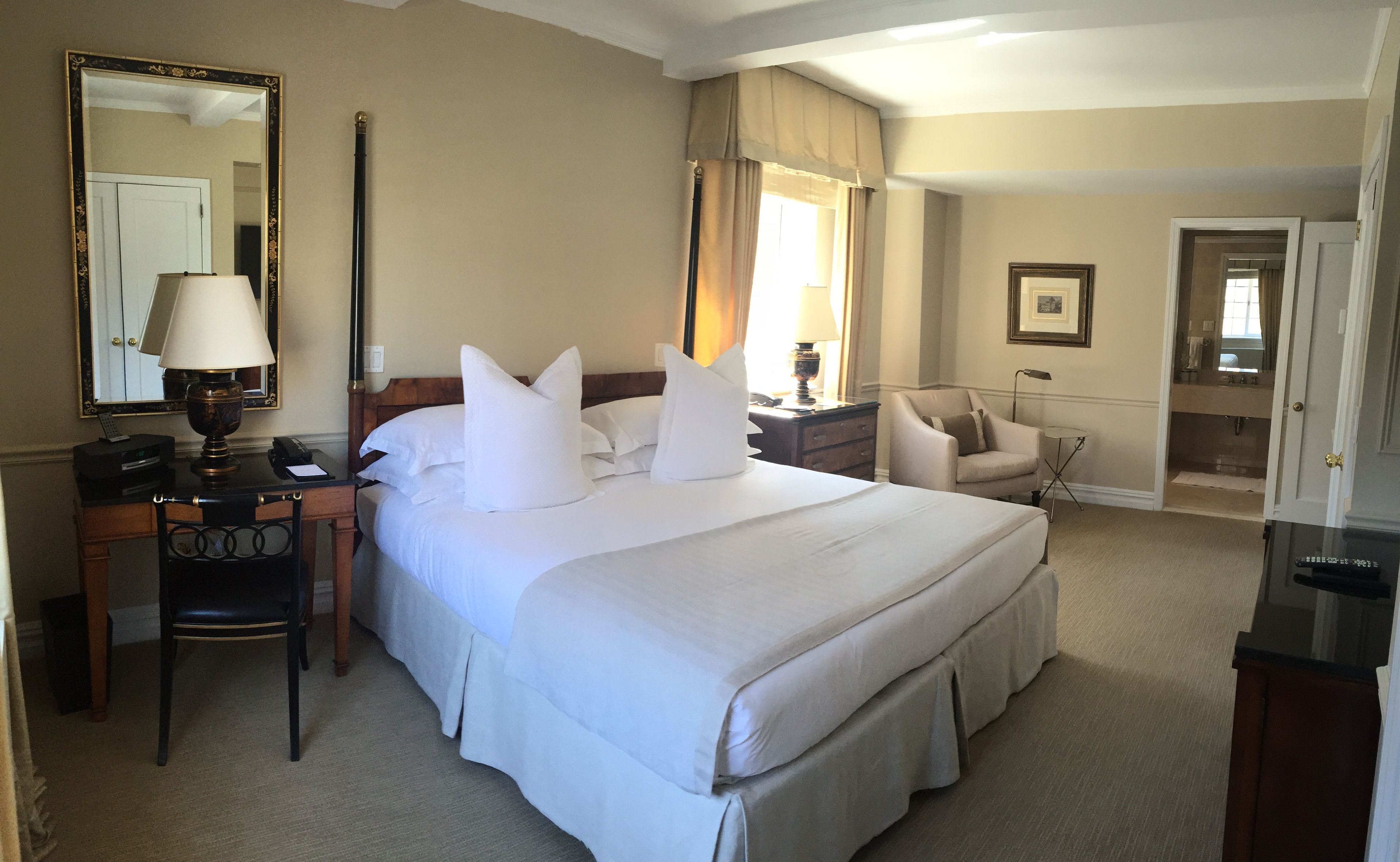 A hotel room with a large bed, a desk with a lamp, a chair, a bedside table, and a mirror.