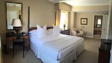A hotel room with a large bed, a desk with a lamp, a chair, a bedside table, and a mirror.