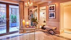 Um corredor bem iluminado com uma mesa de vidro exibindo livros e velas, um banco e uma porta que dá para um quarto com quadros.