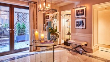 A well-lit hallway with a glass table displaying books and candles, a bench, and a door leading to a room with framed pictures.