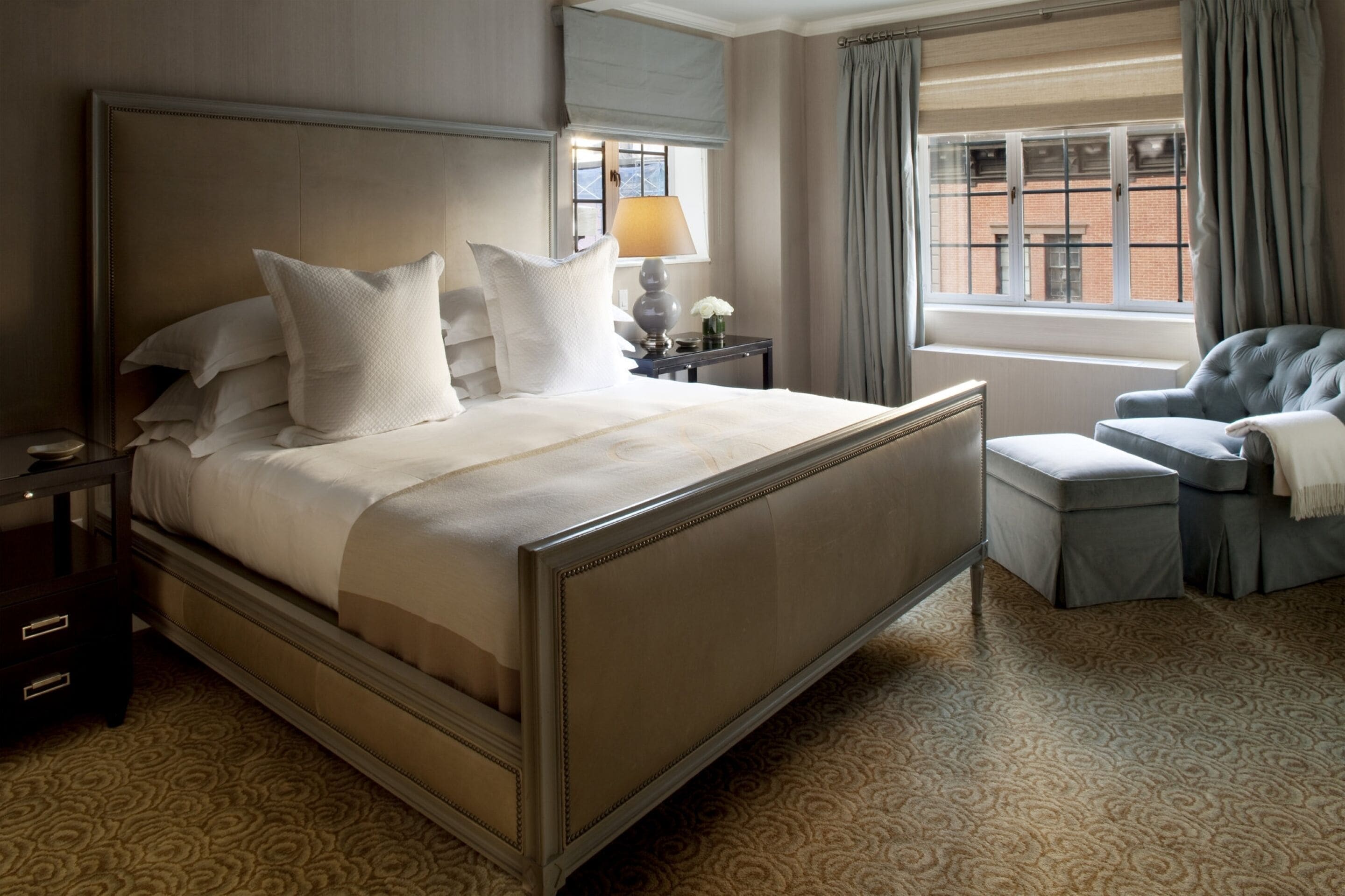Ein ordentlich eingerichtetes Hotelzimmer mit einem großen Bett, einem Sessel, einem Nachttisch, einer Lampe und einem Fenster mit Vorhängen.