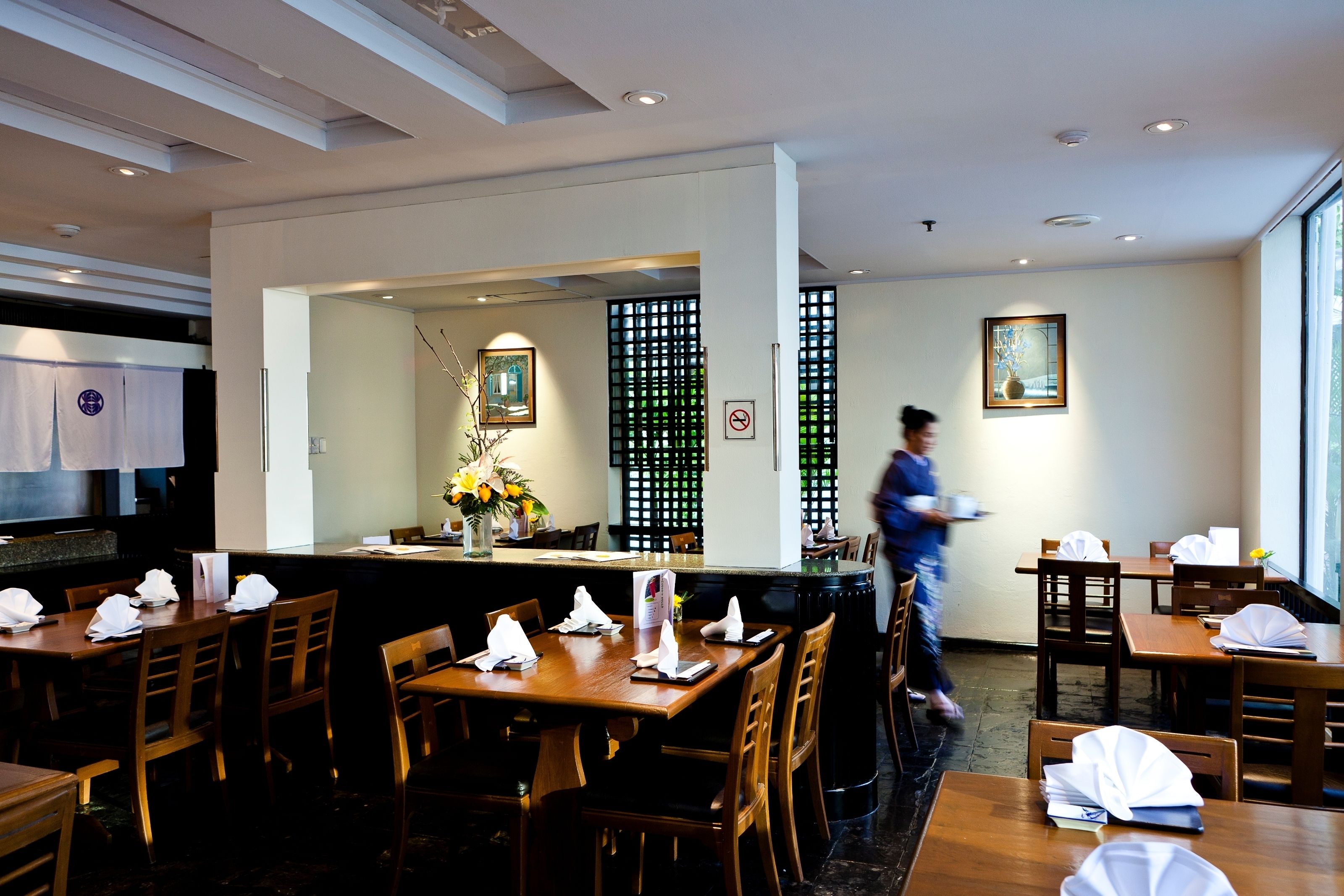 A restaurant with wooden tables and chairs, a counter, and a person walking in the background.