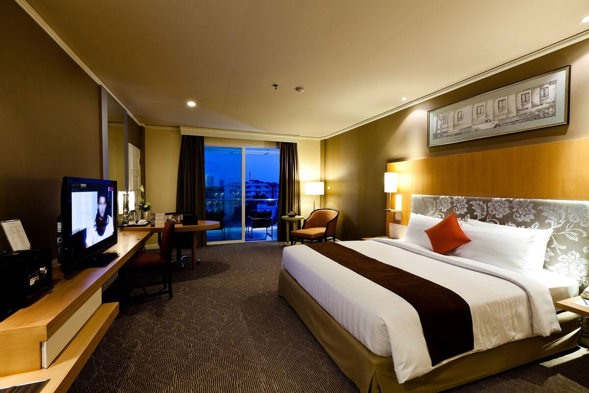 A hotel room with a large bed, a desk with a computer, a chair, a TV, and a view of the city at dusk.