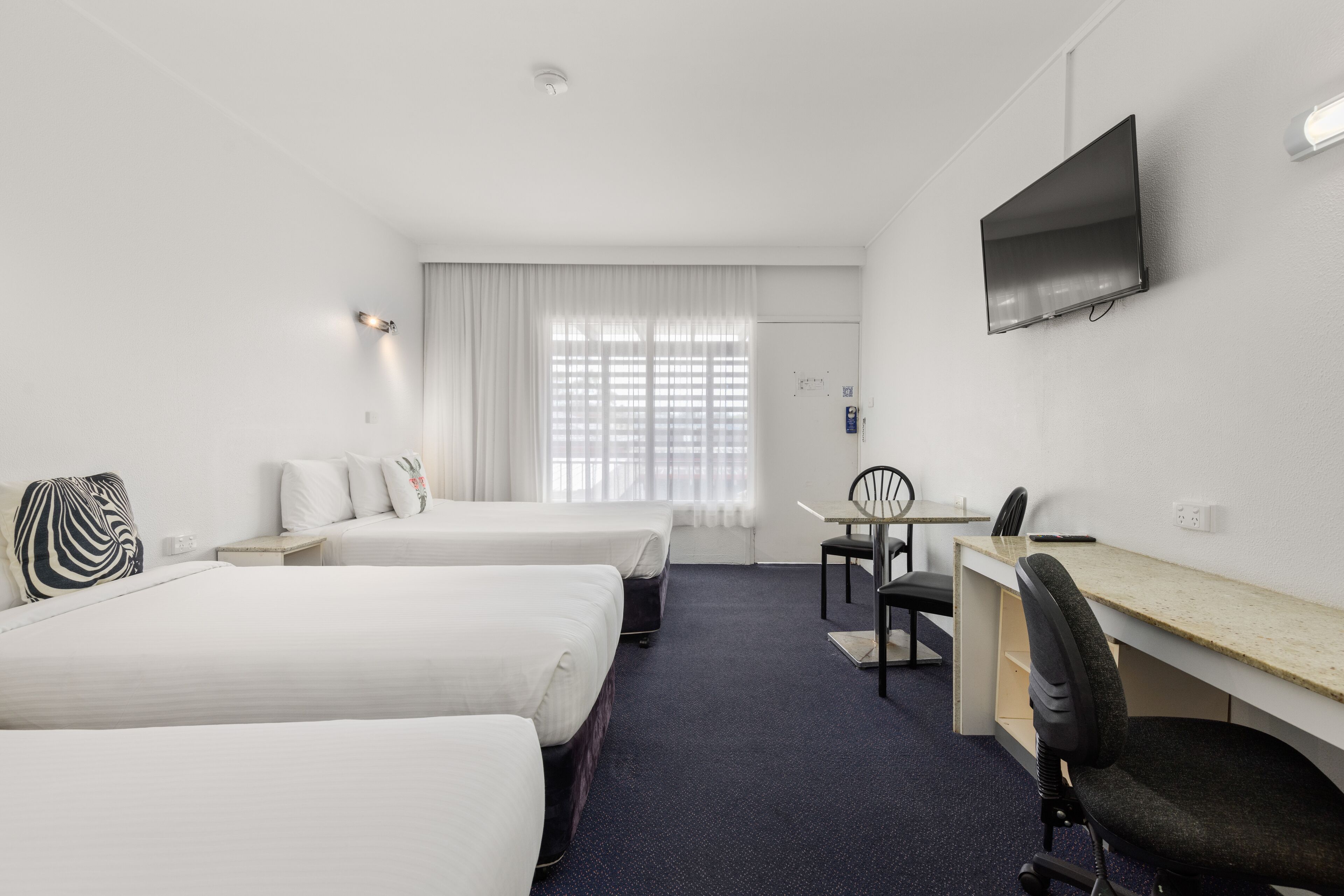 Quarto de hotel com duas camas, uma escrivaninha, uma cadeira, televisão e janela com cortinas.