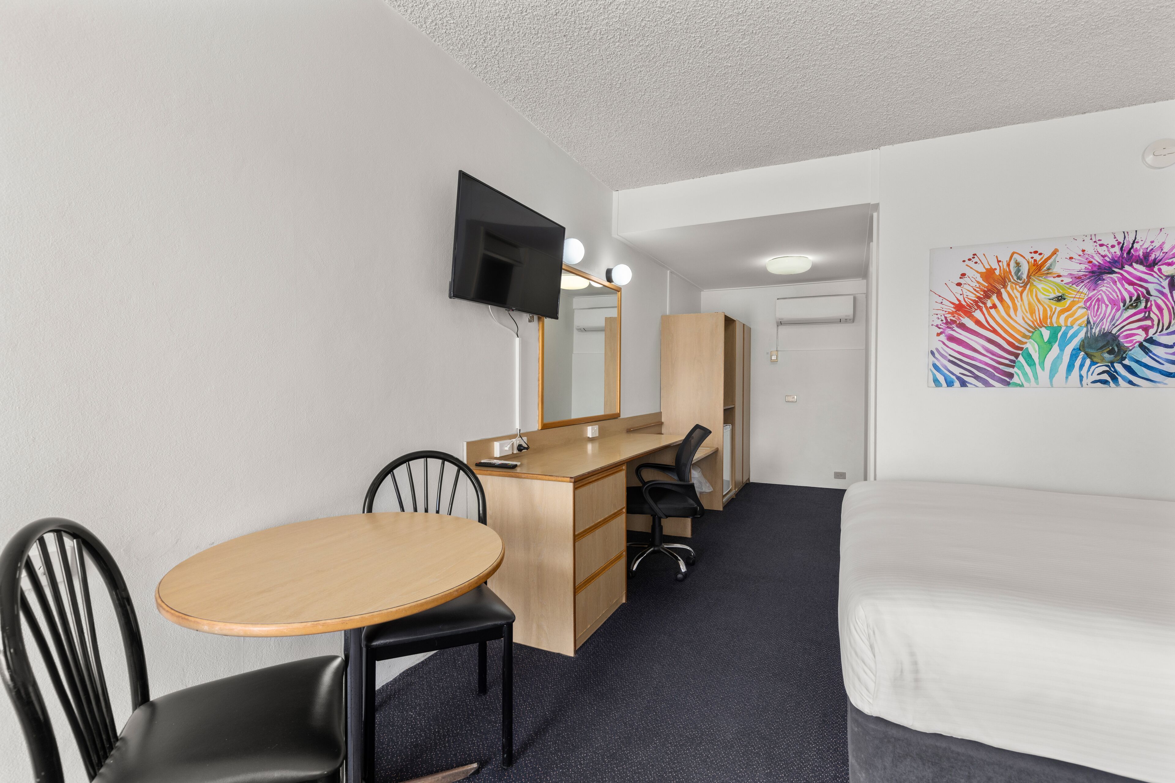 Habitación de hotel con cama, escritorio con silla, televisor montado en la pared y espejo.