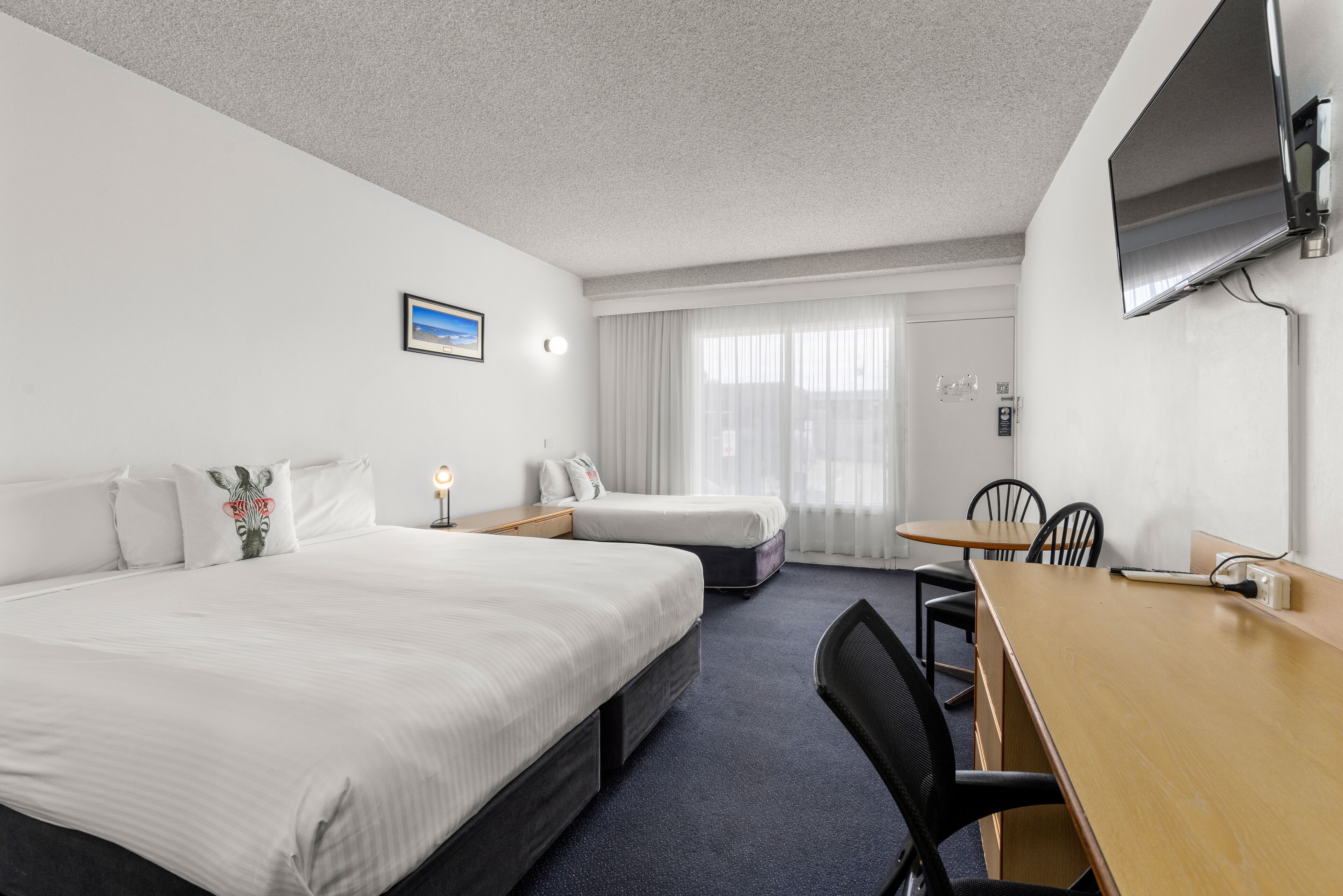 A hotel room with two beds, a desk, chairs, a TV, and a window with curtains.