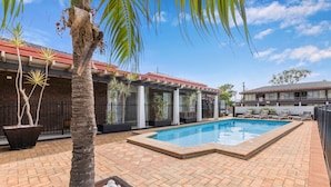 A swimming pool with a tiled deck, palm trees, and a building with a red roof in the background. - Zebra Motel   (Coffs Harbour)