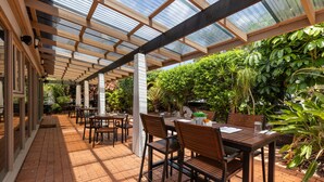 A covered patio area with wooden tables and chairs, surrounded by lush greenery and a glass ceiling structure. - Zebra Motel   (Coffs Harbour)