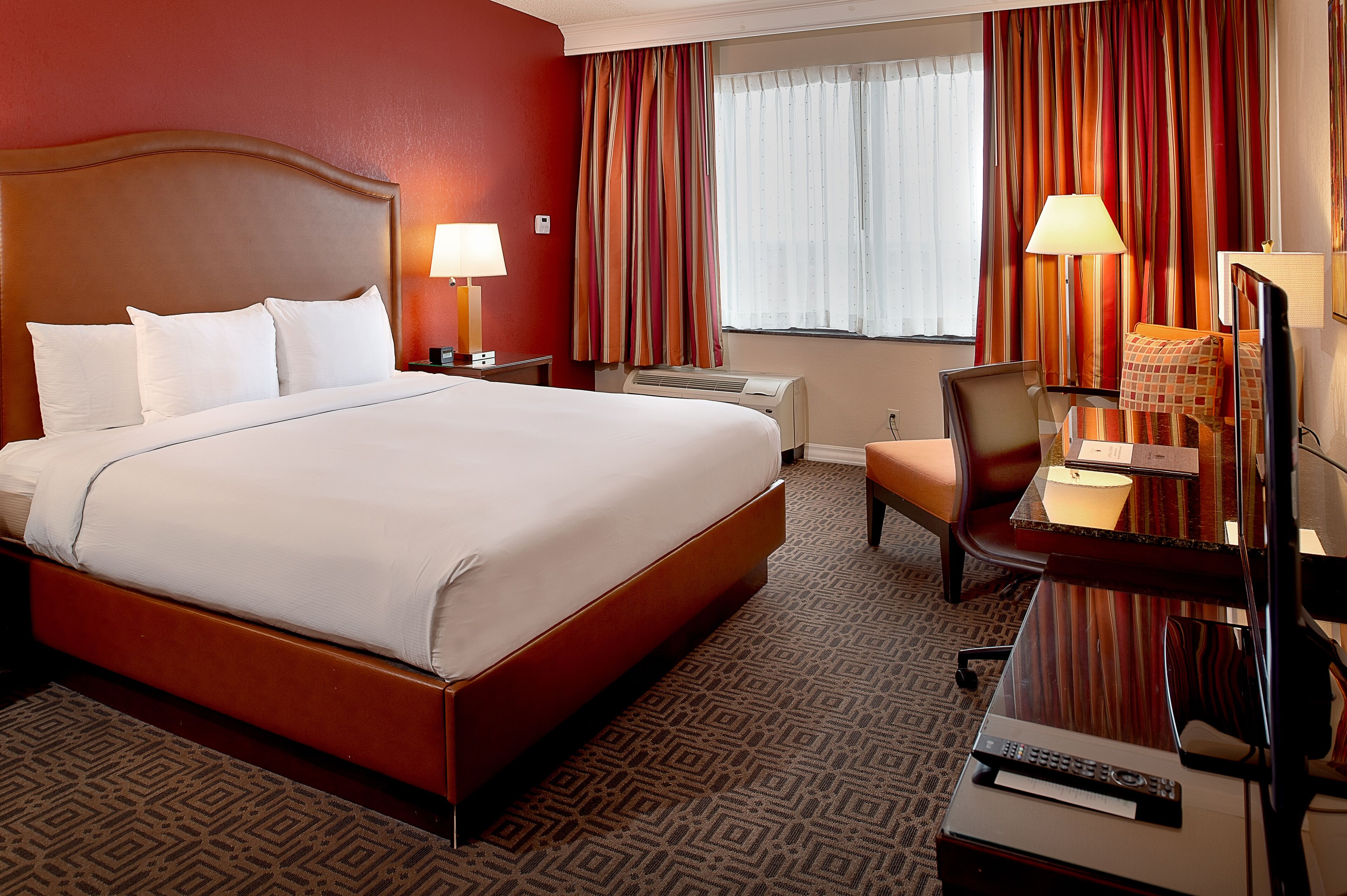 Quarto de hotel com uma cama grande, uma escrivaninha com cadeira, uma luminária e uma televisão.