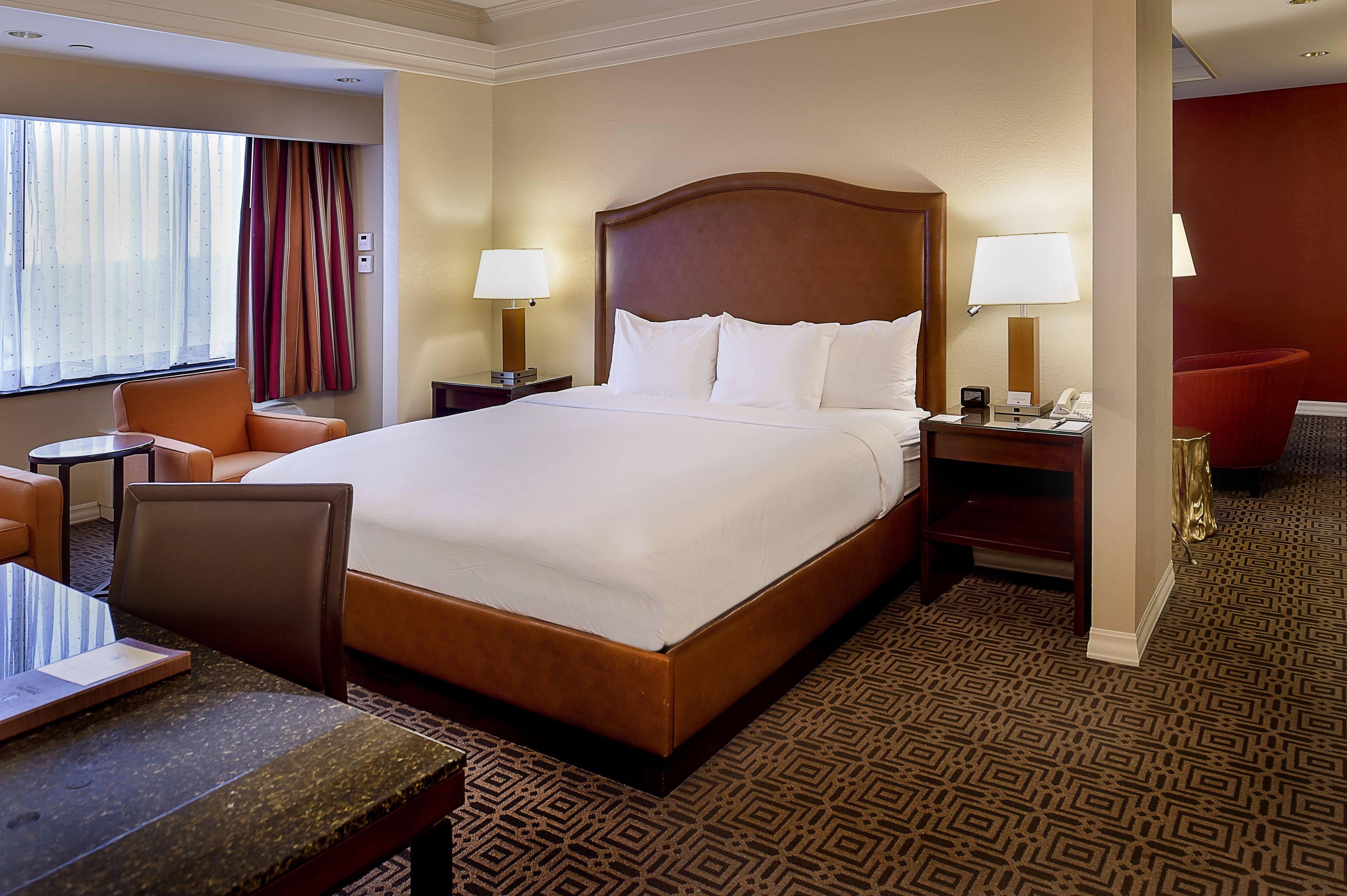Habitación de hotel con una cama grande, dos mesitas de noche con lámparas, una pequeña mesa con una silla y un ventanal con cortinas.