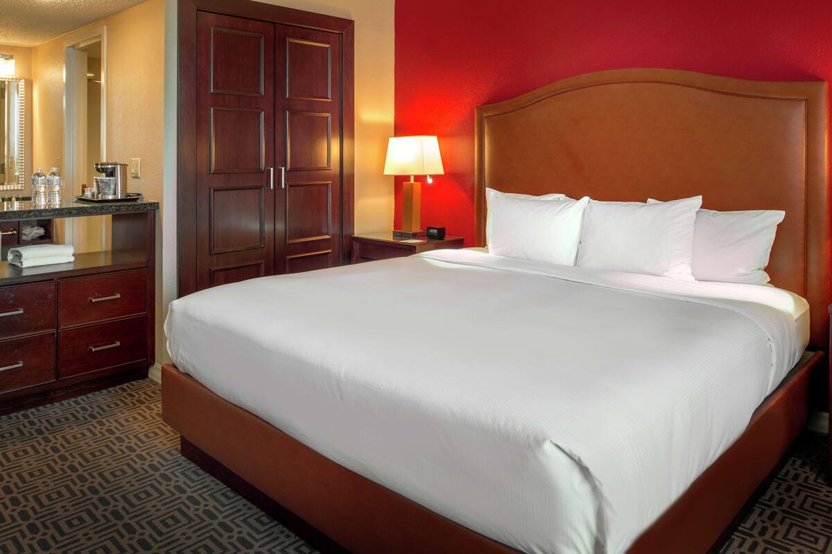 Une chambre d’hôtel avec un grand lit, une tête de lit en bois, une table de chevet avec une lampe, une commode avec un miroir et un mur d’accent rouge.
