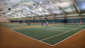 Sisätiloissa sijaitseva tenniskenttä, jossa on useita kenttiä, verkko ja pelaajia.