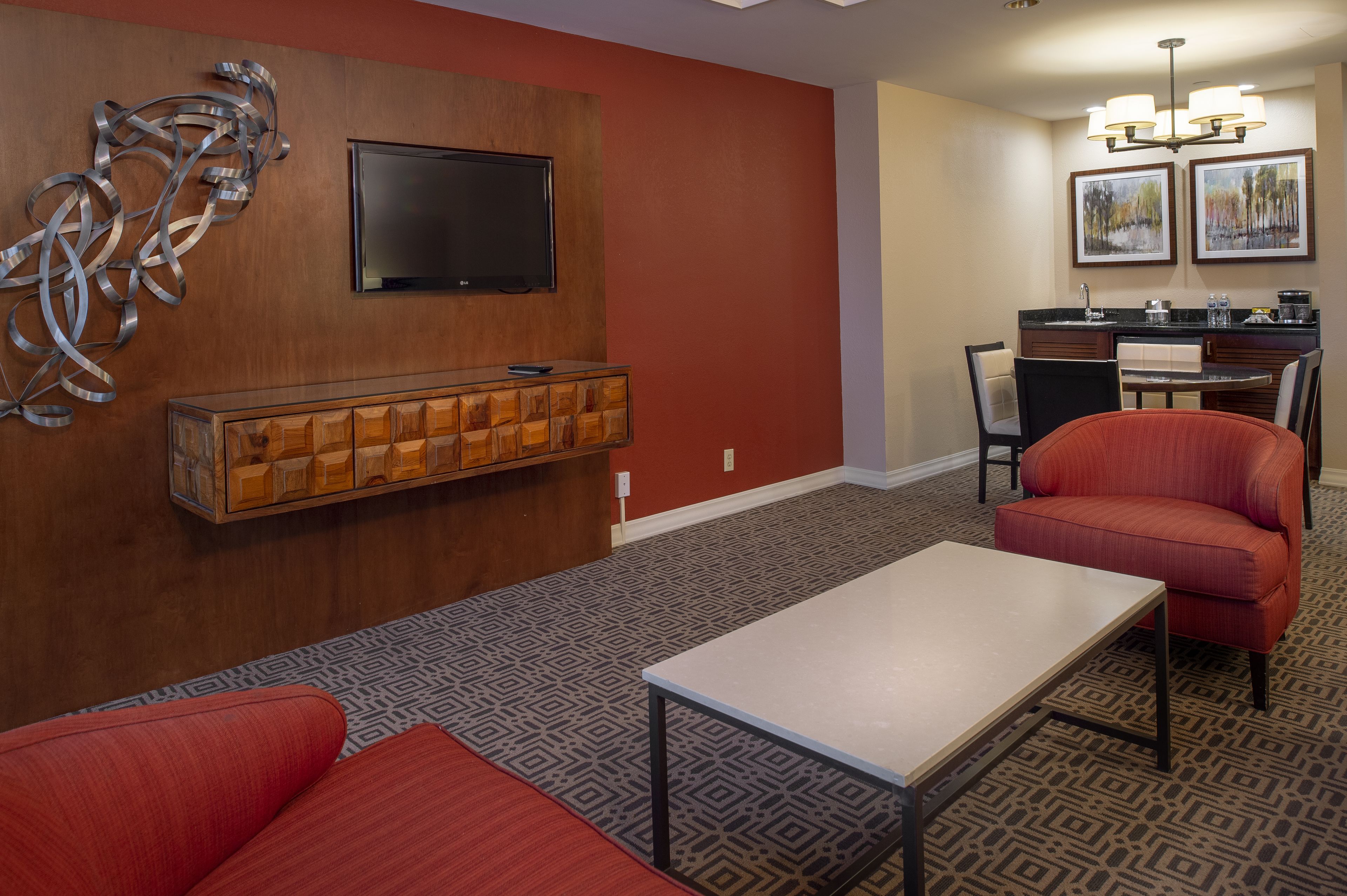A hotel room with a TV mounted on the wall, a wooden cabinet below it, a dining area with a table and chairs, and a red sofa.