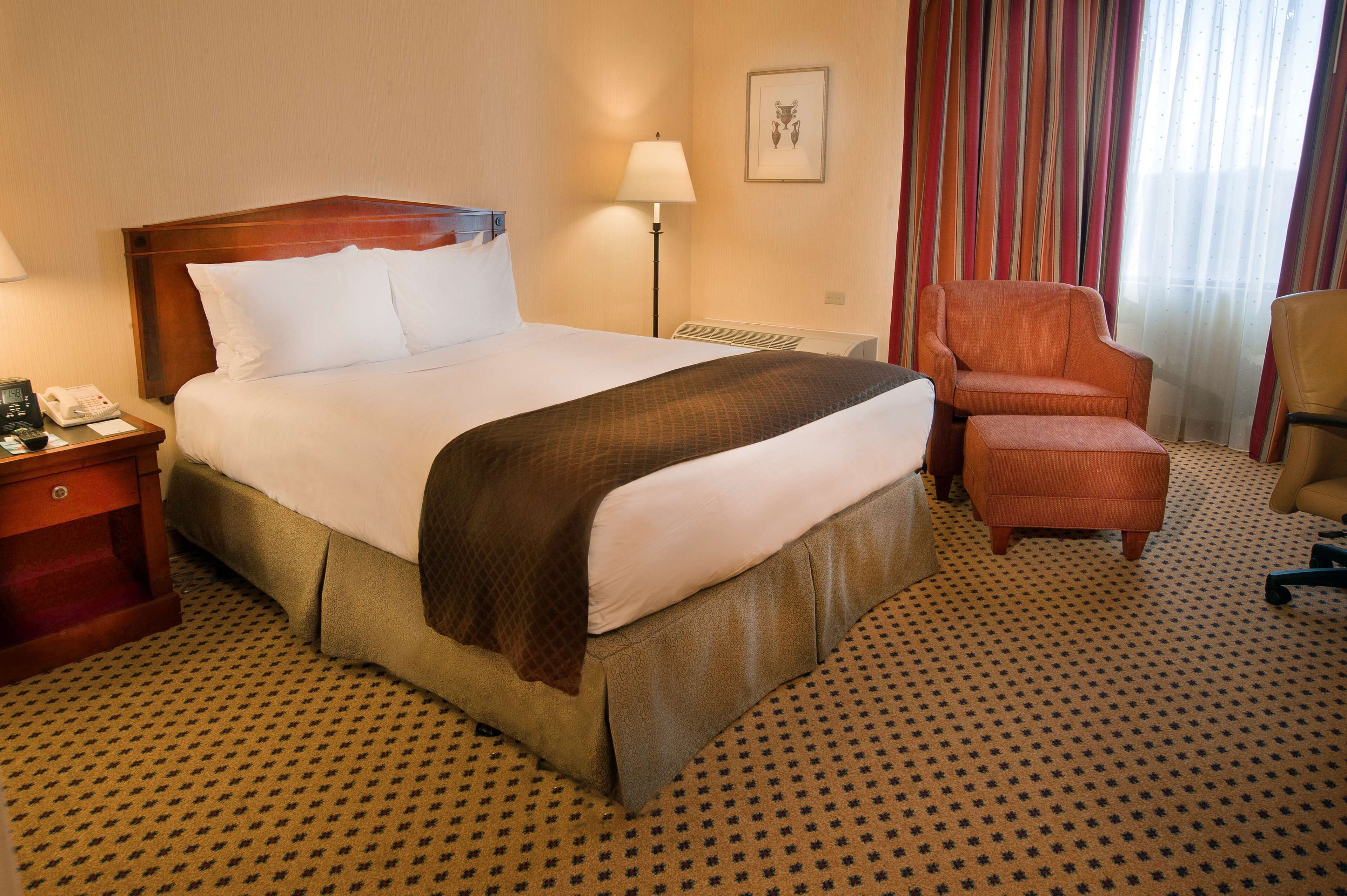 Une chambre d’hôtel avec un grand lit, une table de chevet avec un téléphone, une lampe, une chaise et un sol recouvert de moquette.