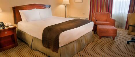 Une chambre d’hôtel avec un grand lit, une table de chevet avec un téléphone, une lampe, une chaise et un sol recouvert de moquette.
