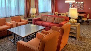 Hotellihuone, jossa on oranssit huonekalut, lasinen sohvapöytä, ruokailutila pöydän ja tuolien kera sekä kuviollinen matto.