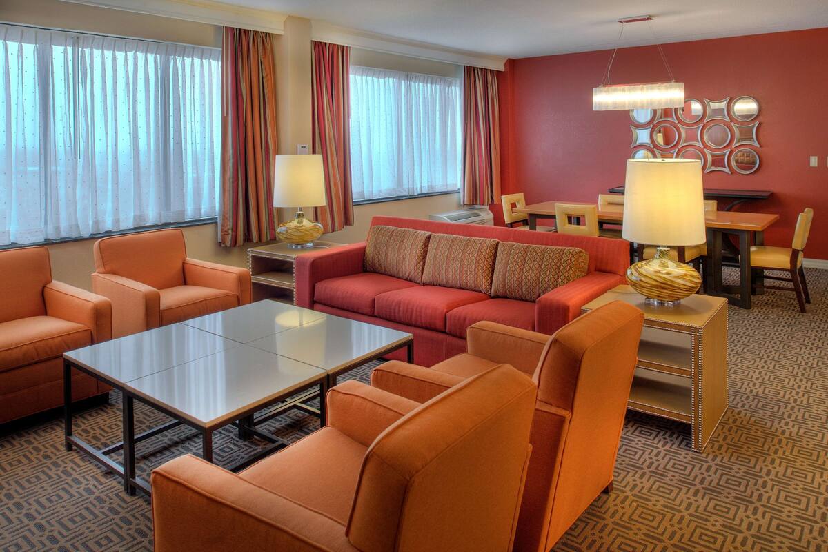 Une chambre d’hôtel avec des meubles de couleur orange, une table basse en verre, un coin repas avec une table et des chaises, et un tapis à motifs.