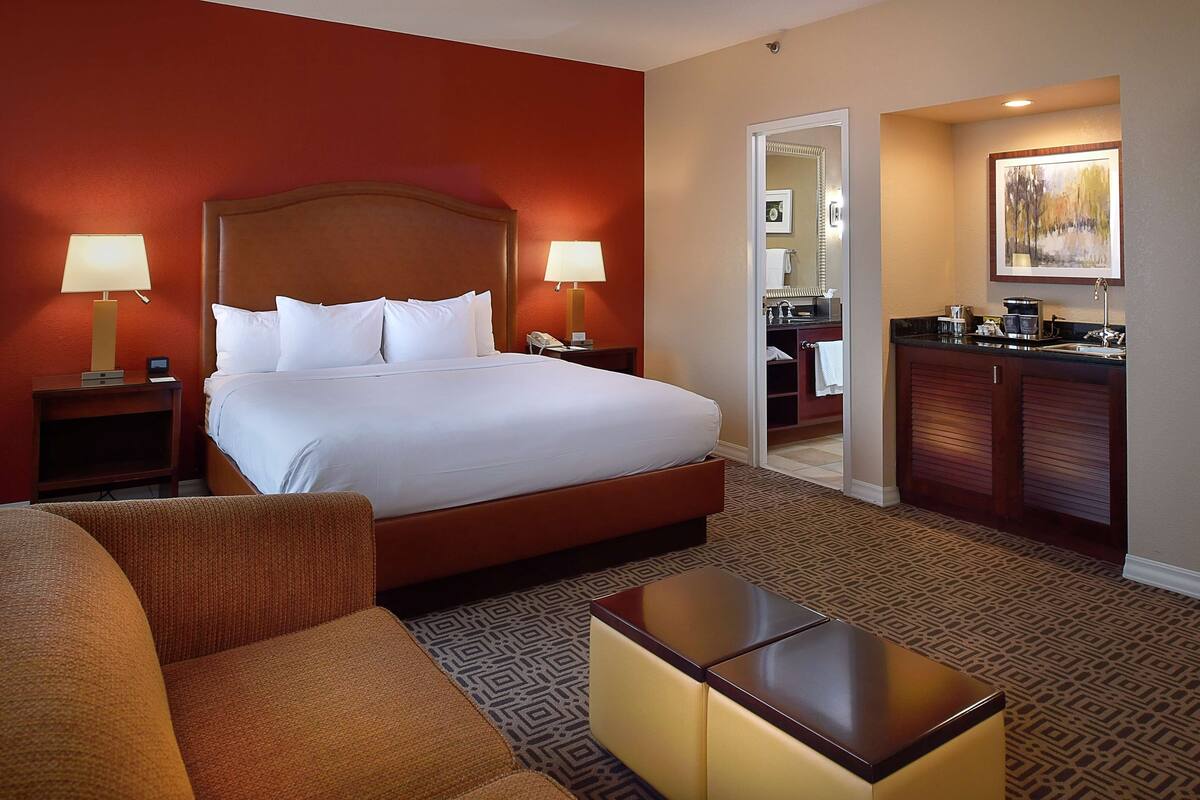Une chambre d’hôtel avec un grand lit, deux tables de chevet avec des lampes, un canapé, une petite table et un coin cuisine.