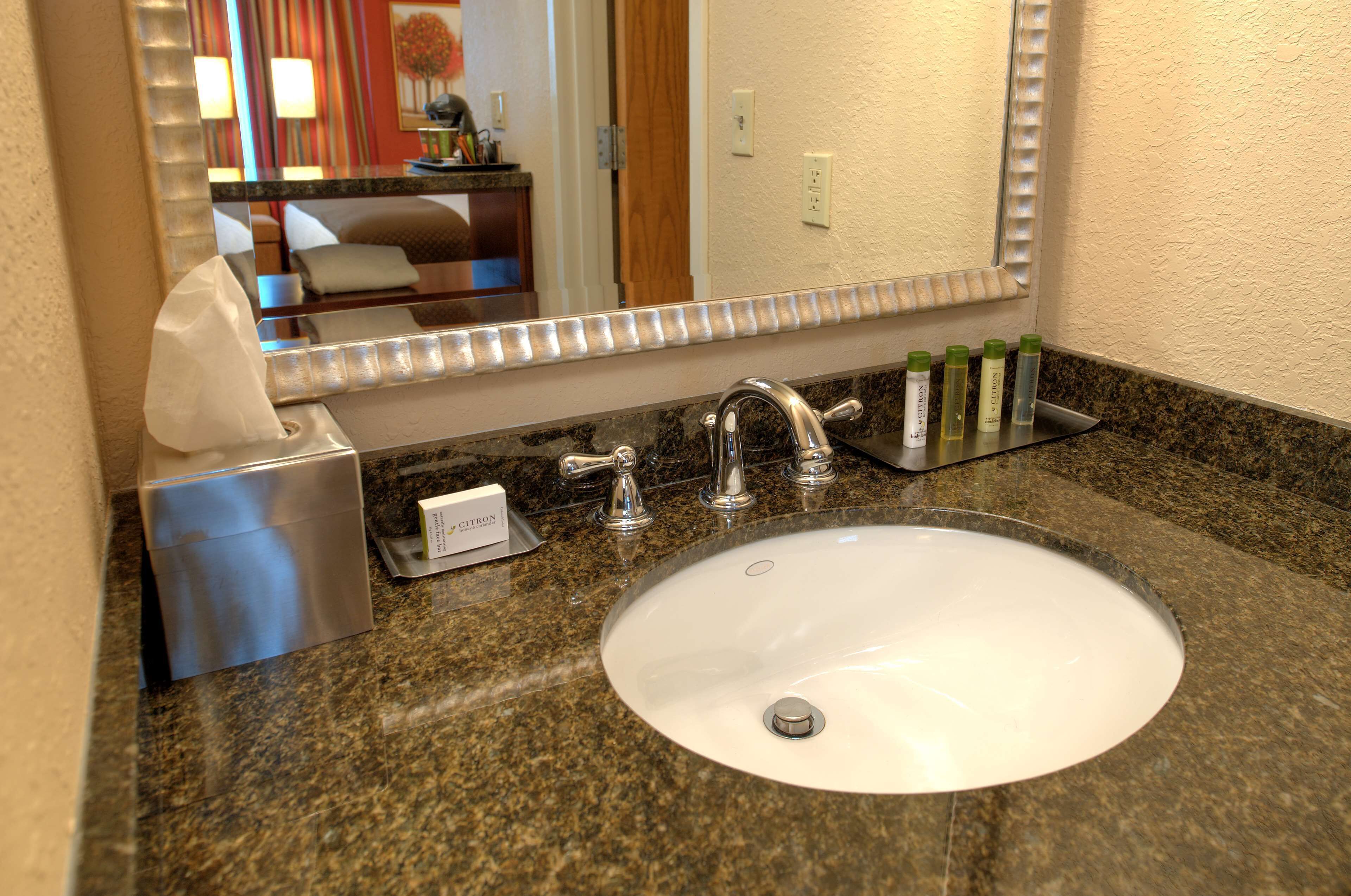 A hotel bathroom with a granite countertop, a white sink, a mirror, and toiletries.